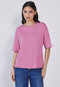 Giovane donna che indossa una maglietta rosa larga a maniche corte e jeans blu, in piedi contro uno sfondo grigio chiaro uniforme.