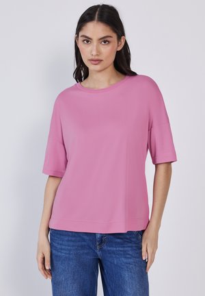 Jeune femme portant un haut rose à manches courtes ample et un jean bleu, se tenant devant un fond gris clair uni.