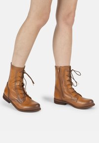 Botas de tobillo de cuero marrón claro con punta redonda, cordones en el frente y cremallera lateral. Presentan una textura lisa y un tacón bajo y apilado.