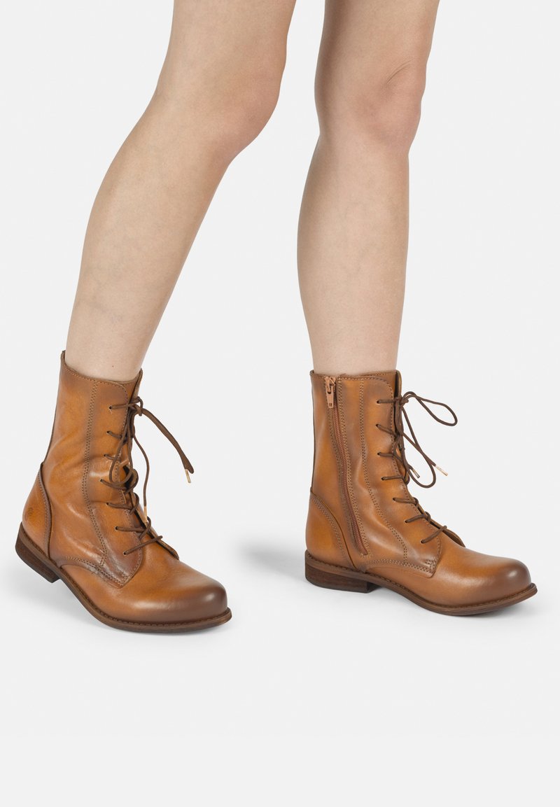 Botas de tobillo de cuero marrón claro con punta redonda, cordones en el frente y cremallera lateral. Presentan una textura lisa y un tacón bajo y apilado.