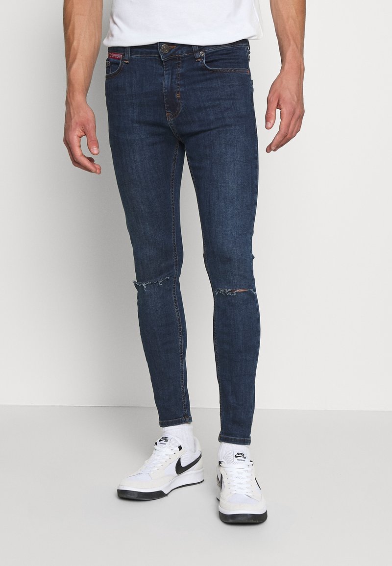 Jeans ajustados de color azul oscuro con detalles de desgaste en las rodillas, fabricados en material de mezclilla. Combinados con zapatillas blancas, exhibiendo un estilo casual.