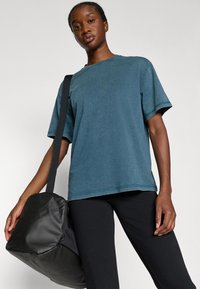 Mėlynos spalvos medvilnės marškiniai su laisvu siluetu, trumpomis rankovėmis ir apvaliu iškirpimu, derinami su juodomis kelnėmis ir juoda rankine su petnešomis.