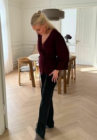 Pulover burgundy cu mâneci lungi și curea legată, purtat împreună cu pantaloni negri evazați. Podea de lemn deschis și decor în tonuri neutre în fundal.