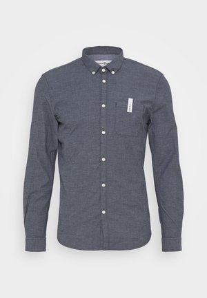 Chemise pour homme à manches longues, fermée par des boutons, de couleur gris foncé, avec une poche poitrine ornée d'une petite étiquette blanche, sur un fond uni.