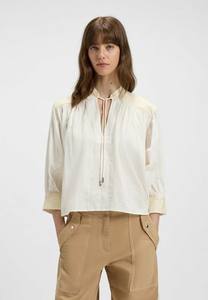 Blusa bianca con colletto alla cinese, chiusura con fiocco al collo, spalle ricamate e maniche a 3/4. Tessuto morbido e strutturato; vestibilità leggermente larga.