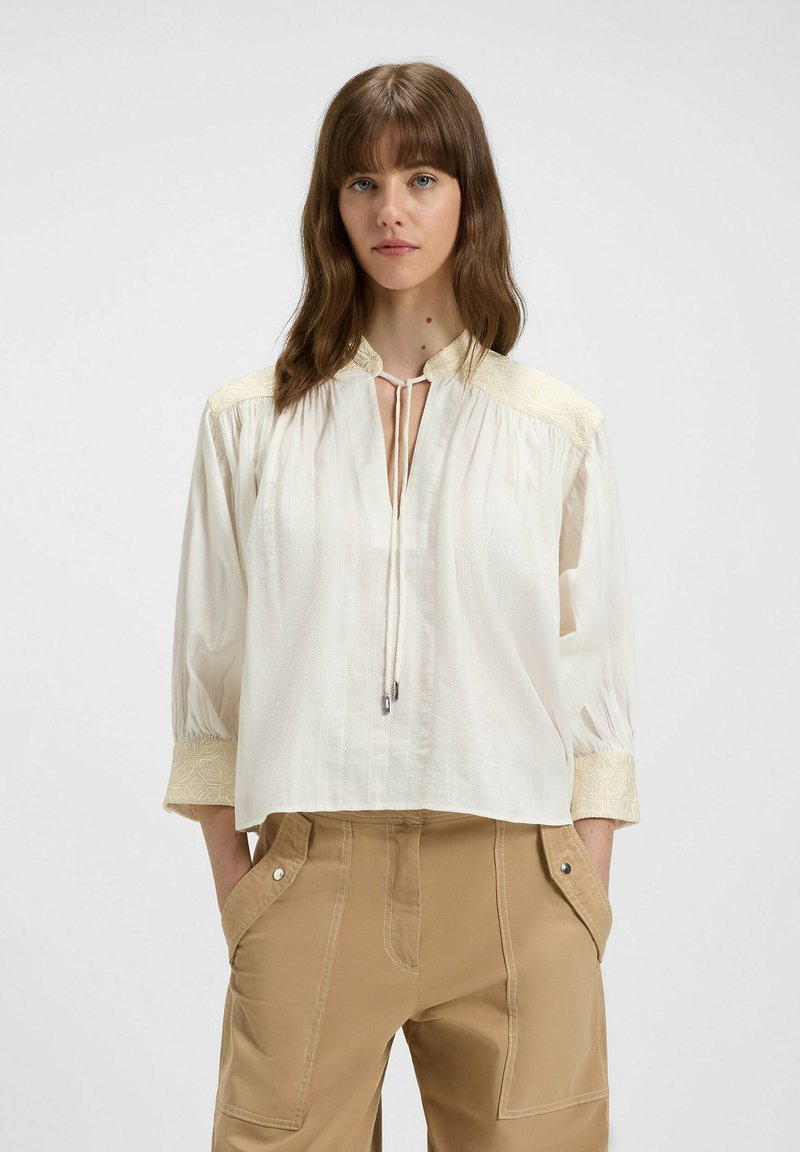 White blouse with a mandarin collar, tie neck closure, embroidered shoulders, and 3/4 sleeves. Soft, textured fabric; slightly loose fit.