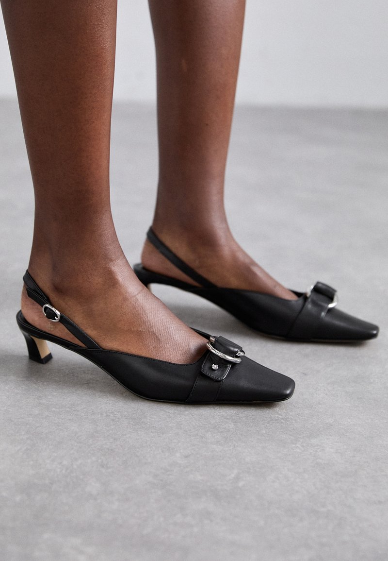 Svarta slingbackklackar med spetsiga tår, lädermaterial, liten blockklack, silverdetalj med spänne på ovansidan, elegant design.
