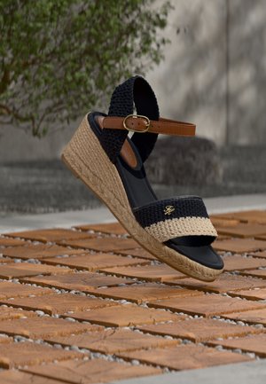 Sandalia de cuña tejida en negro y beige con correa marrón en el tobillo y hebilla dorada, flotando sobre baldosas de terracota al aire libre cerca de follaje verde.