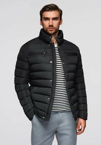 Černá péřová bunda se stoj�áčkem, horizontálním prošíváním, dvěma zipovými kapsami na hrudi a knoflíkovým zapínáním. Nositá přes pruhovanou košili.