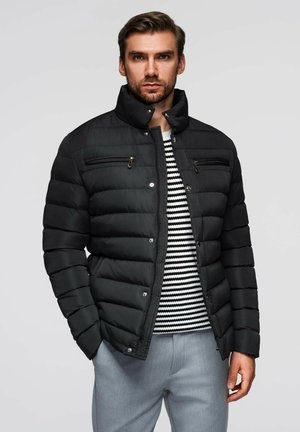 Zwarte pufferjack met een staande kraag, horizontale stikking, twee ritszakken op de borst en knoopsluitingen. Draagt over een gestreept shirt.