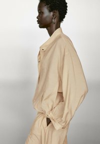 Beige oversized jacka med avslappnad passform, med tryckknappar och sidofickor, tillverkad av lätt tyg med en slät textur.