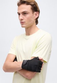 Νέος άντρας με ανοιχτά μαλλιά, που φοράει ένα απαλό κίτρινο t-shirt και μαύρα γάντια χωρίς δάχτυλα, με σταυρωμένα τα χέρια, κοιτάζει προς τα πλάγια πάνω σε ένα απλό φόντο.