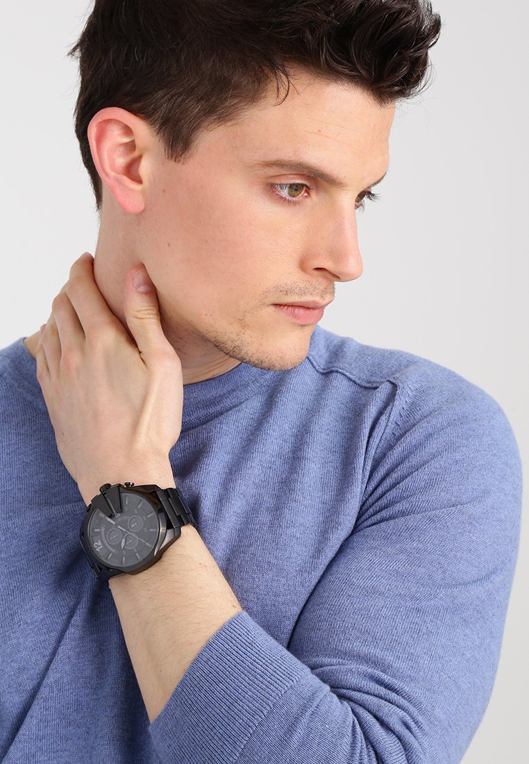 Czarny analogowy zegarek z wyraźną tarczą i fakturowanym paskiem, noszony na nadgarstku osoby w jasnoniebieskim swetrze. Zbliżenie.