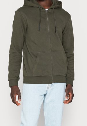 Mann trägt olivgrünen Hoodie mit Reißverschluss und Fronttaschen sowie hellblaue Jeans, steht vor einem schlichten Hintergrund.