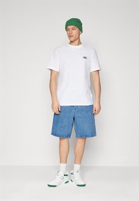 White cotton t-shirt with a small black logo, paired with blue denim shorts and white sneakers with green accents. Green knit beanie.