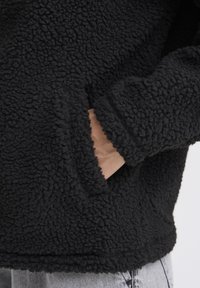 Mustast fliisist pullover, millel on tekstuuriga pehme pind. Omab esipocketit ja soonikkoesid. Alläär on sirge ja lihtne.