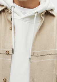 Veste chemise beige à motif avec des boutons, superposée à un sweatshirt à capuche blanc. La veste présente un tissage texturé et des accents gris.