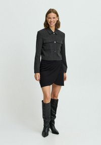 Veste grise courte avec deux poches avant, fermeture à boutons, jupe noire enveloppante et bottes noires montantes avec une texture lisse.