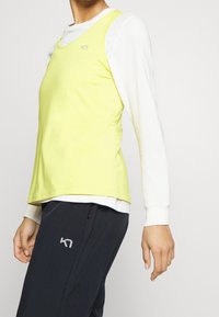 Ärmlös gul linne med en slät textur, lager-på-lager över en långärmad vit skjorta, ihop med mörka byxor med en logotyp.