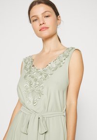 Vestido verde claro com decote redondo, bordados florais em prata e um laço de tecido na cintura. Textura suave e fluida.