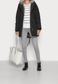 Person som bär en svart parkas med fuskpäls, en vit randig tröja, grå skinny jeans och svarta ankelboots, håller en stor vit tote-väska, står inomhus.