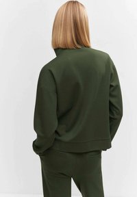 Sudadera verde oliva con cuello alto, corte ancho, hombros caídos y puños acanalados. Textura suave con diseño minimalista.