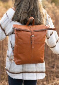 Una persona che indossa una giacca a quadretti bianca e pantaloni scuri porta uno grande zaino di pelle marrone all'aperto in un'area erbosa secca.