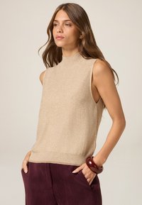 Top de punto sin mangas color beige con cuello alto, textura suave y acentos brillantes, combinado con pantalones de pana color púrpura oscuro y una pulsera marrón.