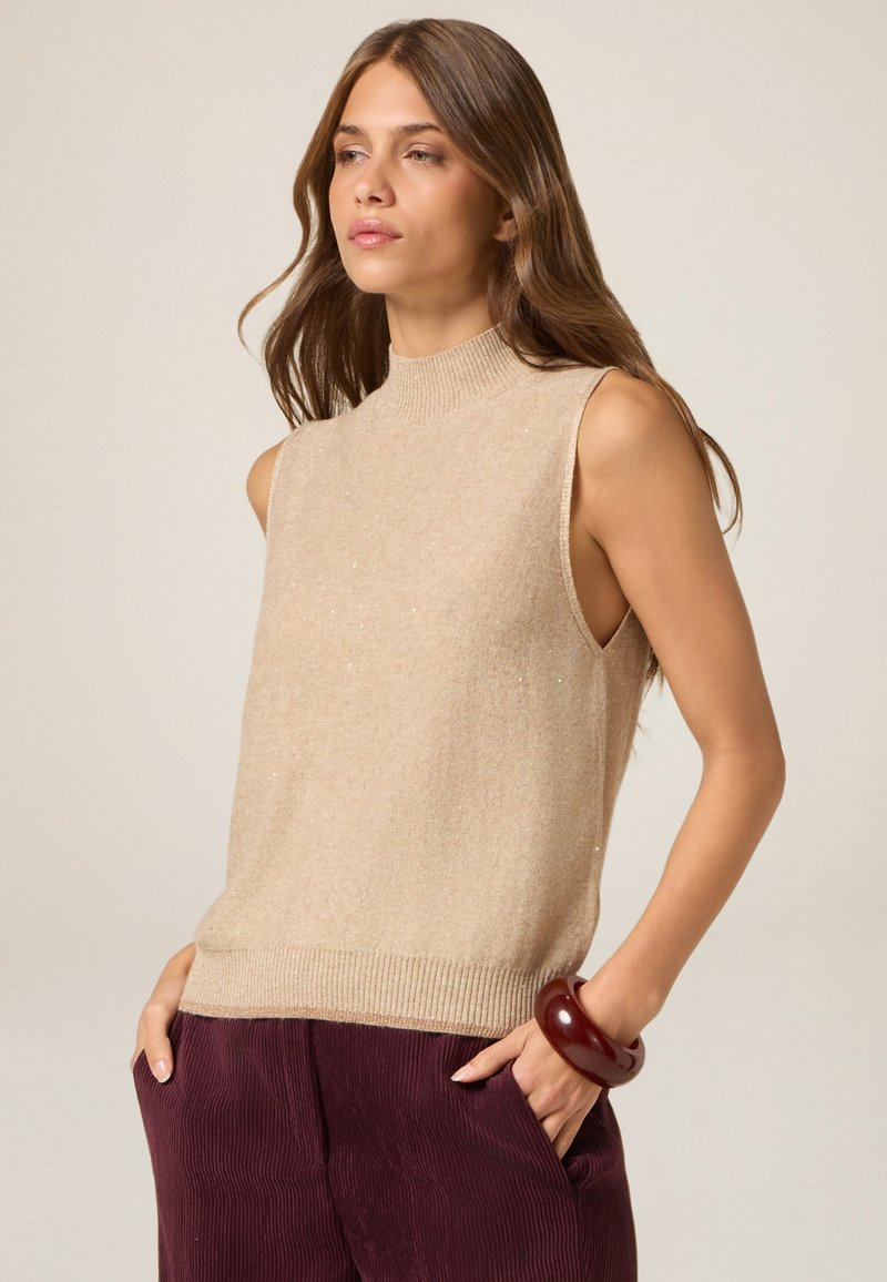 Top de punto sin mangas color beige con cuello alto, textura suave y acentos brillantes, combinado con pantalones de pana color púrpura oscuro y una pulsera marrón.