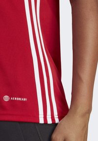 Rode sportieve shirt met witte verticale strepen aan de zijkant. Gemaakt van ademend materiaal, met het logo "AEROREADY" nabij de zoom.
