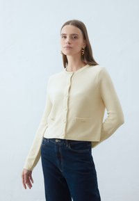 Kremowy, dzianinowy kardigan z okrągłym dekoltem, zapięcie na guziki z przodu i dwiema kieszeniami. Połączony z ciemnoniebieskimi dżinsami.