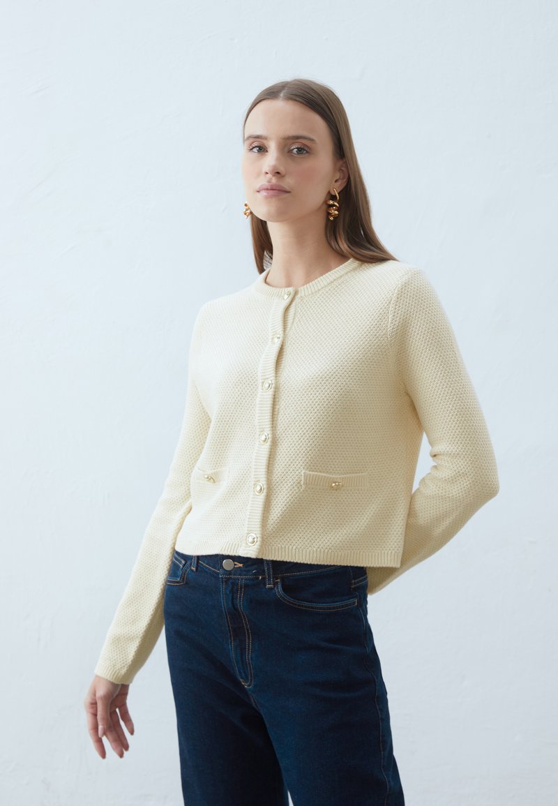 Kremowy, dzianinowy kardigan z okrągłym dekoltem, zapięcie na guziki z przodu i dwiema kieszeniami. Połączony z ciemnoniebieskimi dżinsami.