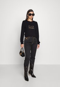 Zwarte sweatshirt met gouden EA7-logo, gecombineerd met zwarte broek met gouden stiksels. Zwarte stiletto laarzen en een kleine zwarte handtas.