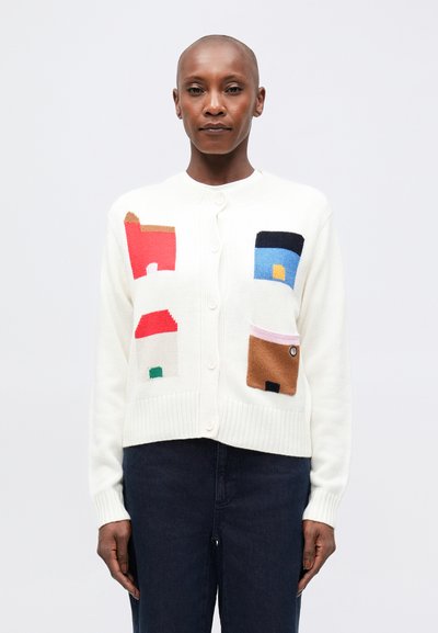 Cardigan crème avec des motifs de maisons colorées, présentant des motifs rouges, bleus et verts. Fermeture à boutons sur le devant, poignets et ourlet côtelés. Tissu tricoté texturé.
