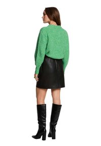 Pullover verde a maglia con motivo testurizzato, abbinato a una mini gonna in pelle nera. Stivali neri con tacco al ginocchio completano l'outfit.
