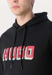 Juodas bliuzonas su dideliu, ryškiu "HUGO" logotipu raudona ir mėlyna spalvomis. Medžiaga atrodo minkšta su tekstūruotu paviršiumi. Yra reguliuojamos raišteliai.