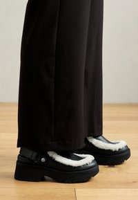 Zwarte platform schoenen hebben een witte bontachtige rand, een ronde neus en een band met een metalen knop. Gecombineerd met zwarte broek op een houten vloer.