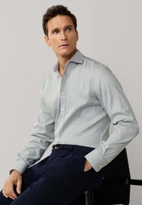 Mann sitzt auf einem Stuhl, trägt ein weiß-grau gestreiftes Langarmshirt und dunkelblaue Hose, schaut nach oben rechts.