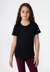 Fiatal lány hosszú göndör hajjal, fekete rövidujjú felsőt és bordó leggingset viselve, egy sima háttér előtt áll.