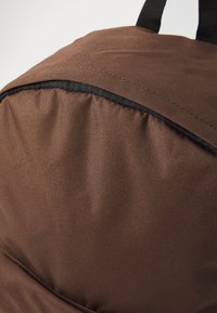 Brązowy plecak z tkaniny o teksturowanej powierzchni, czarnym wykończeniu u góry oraz wyściełanym panelem tylnym dla komfortu. Prosty design, bez wzorów.