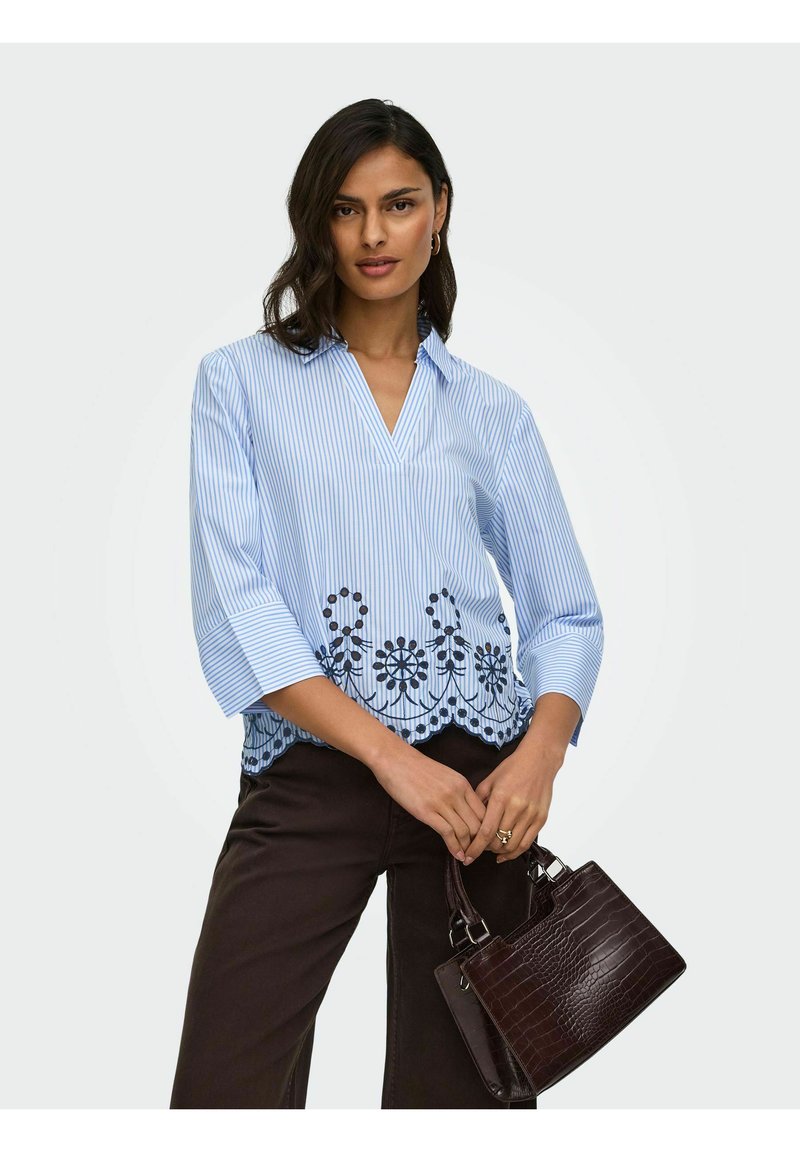 Femme portant un chemisier rayé bleu clair avec broderie florale, tenant un sac à main marron foncé texturé, debout devant un fond uni.