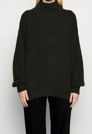 Persoon draagt een losse donker groene gebreide coltrui met geribde manchetten en zoom, gecombineerd met zwarte broek.