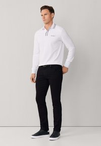 Polo blanco de manga larga con cuello a rayas navy, con un logo en el pecho; combinado con jeans negros de corte ajustado y zapatillas oscuras.