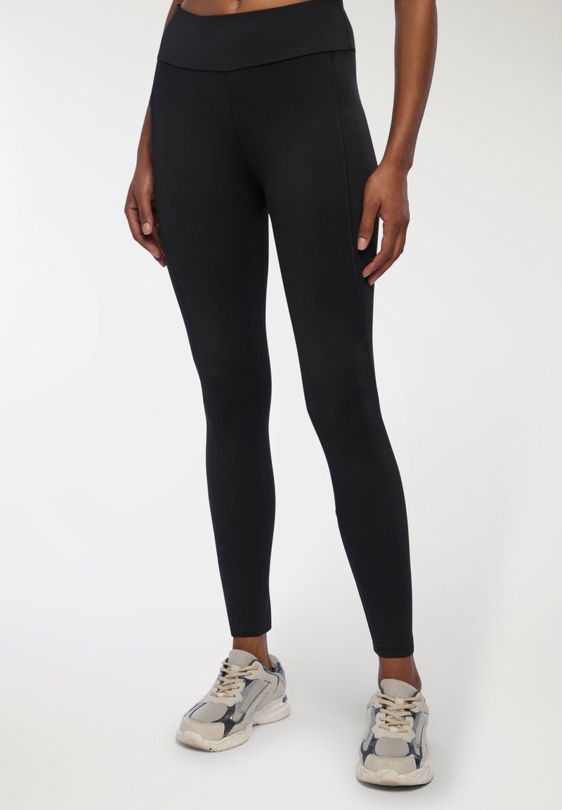 Czarne legginsy o wysokim stanie z gładką teksturą i dopasowanym krojem, w połączeniu z beżowymi i szarymi sneakresami z masywną podeszwą.