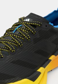 Chaussure de sport noire avec une tige texturée, des lacets jaune vif et noir, et une semelle colorée avec des accents orange.