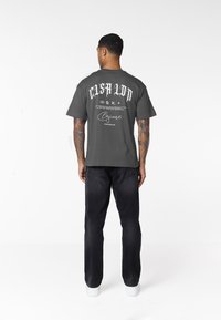 Mann von hinten, trägt ein dunkelgraues T-Shirt mit der Aufschrift "Cash Up", schwarze Jeans, weiße Sneaker und zeigt Armtätowierungen vor weißem Hintergrund.