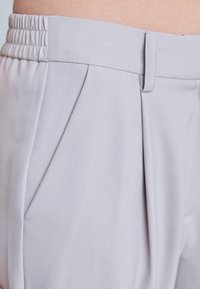 Pantalon gris clair avec une ceinture élastique et des passants de ceinture, présentant des plis et une poche latérale portée à la taille.