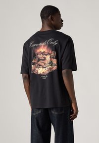 T-shirt de algodão preta com um grande gráfico colorido de comida e bebidas nas costas, com a inscrição "Essentials Only" estilizada acima do gráfico.