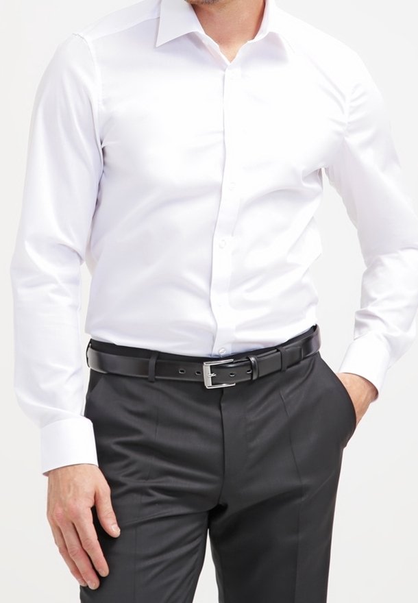 Homme portant une chemise blanche rentrée dans un pantalon de costume noir avec une ceinture en cuir noire, une main dans la poche, sur un fond uni.