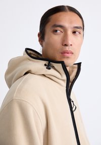 Chaqueta de forro polar beige con una cremallera negra y capucha ajustable. Presenta una textura suave y un diseño minimalista, con un logo en el pecho.